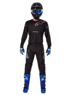 Alpinestars 2025 Racer Graphite Pants, spodnie motocrossowe, czarno-szare, prezentowane na modelu z pasującą czarną koszulką, niebiesko-fioletowymi butami i niebieskimi goglami, kompletny strój wyścigowy off-road z wyraźnym brandingiem Alpinestars