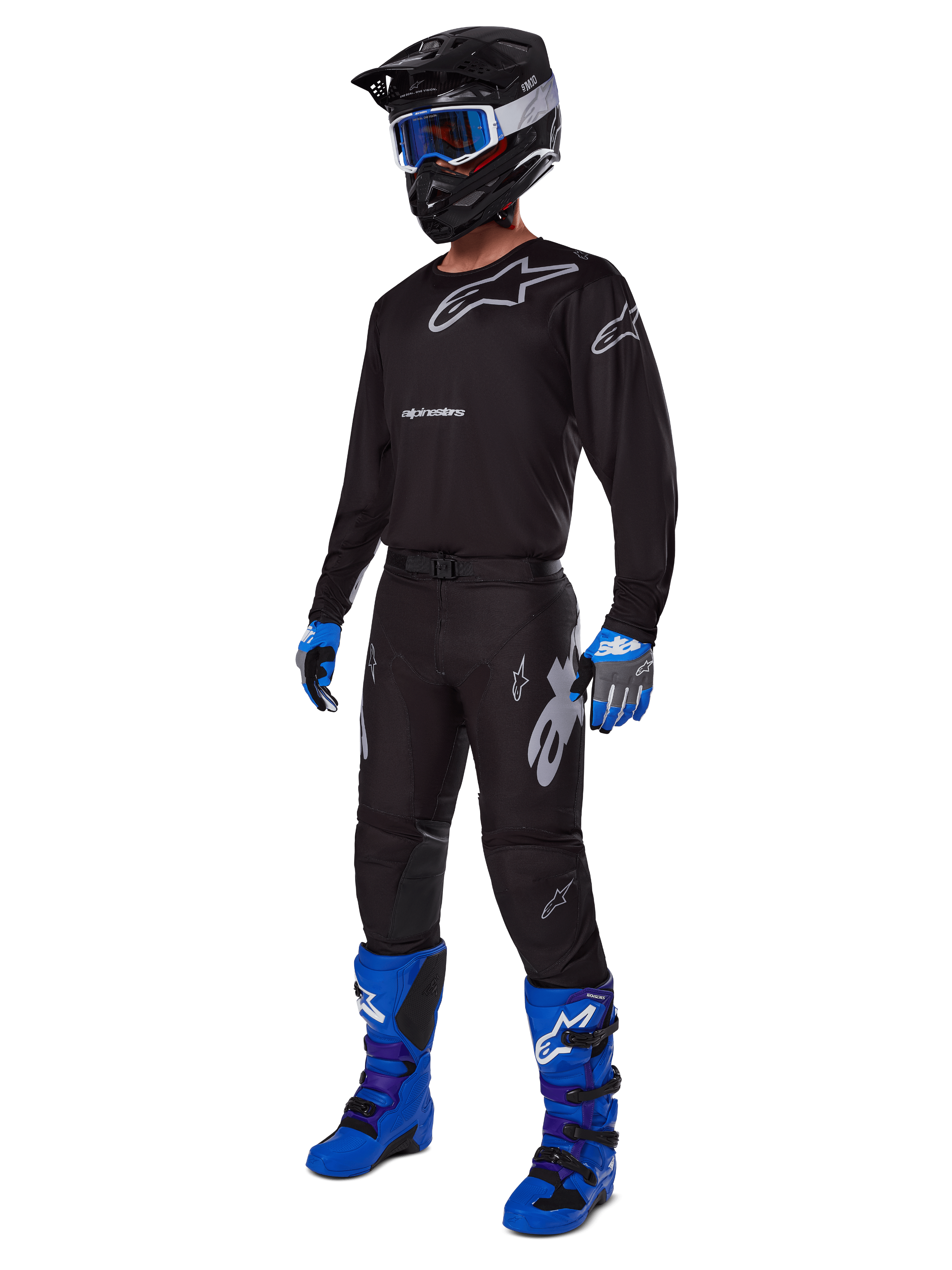 Motocyklista motocrossowy w kompletnym stroju wyścigowym Alpinestars, spodnie Racer Graphite 2025 w kolorze czarno-szarym, pasująca czarna bluza z szarym logo, czarny kask z niebieskimi goglami, jaskrawoniebieskie i fioletowe buty oraz niebiesko-szare rękawice, dopasowany strój do wyścigów terenowych