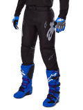 Alpinestars 2025 Racer Graphite Pants, czarne spodnie motocrossowe z białymi i szarymi grafikami, noszone z jasnoniebieskimi butami motocrossowymi i pasującymi niebieskimi rękawicami, wytrzymała tkanina techniczna do jazdy terenowej