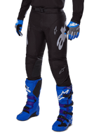 Alpinestars 2025 Racer Graphite Pants, czarne spodnie motocrossowe z białymi i szarymi grafikami, noszone z jasnoniebieskimi butami motocrossowymi i pasującymi niebieskimi rękawicami, wytrzymała tkanina techniczna do jazdy terenowej