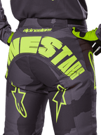 Alpinestars 2025 Racer Hollow Pants, spodnie motocrossowe, kolor ciemne camo i fluorescencyjny żółty, z dużym żółtym logo Alpinestars z tyłu, taktyczny wzór camo z jaskrawymi akcentami, wytrzymała konstrukcja do wyścigów terenowych