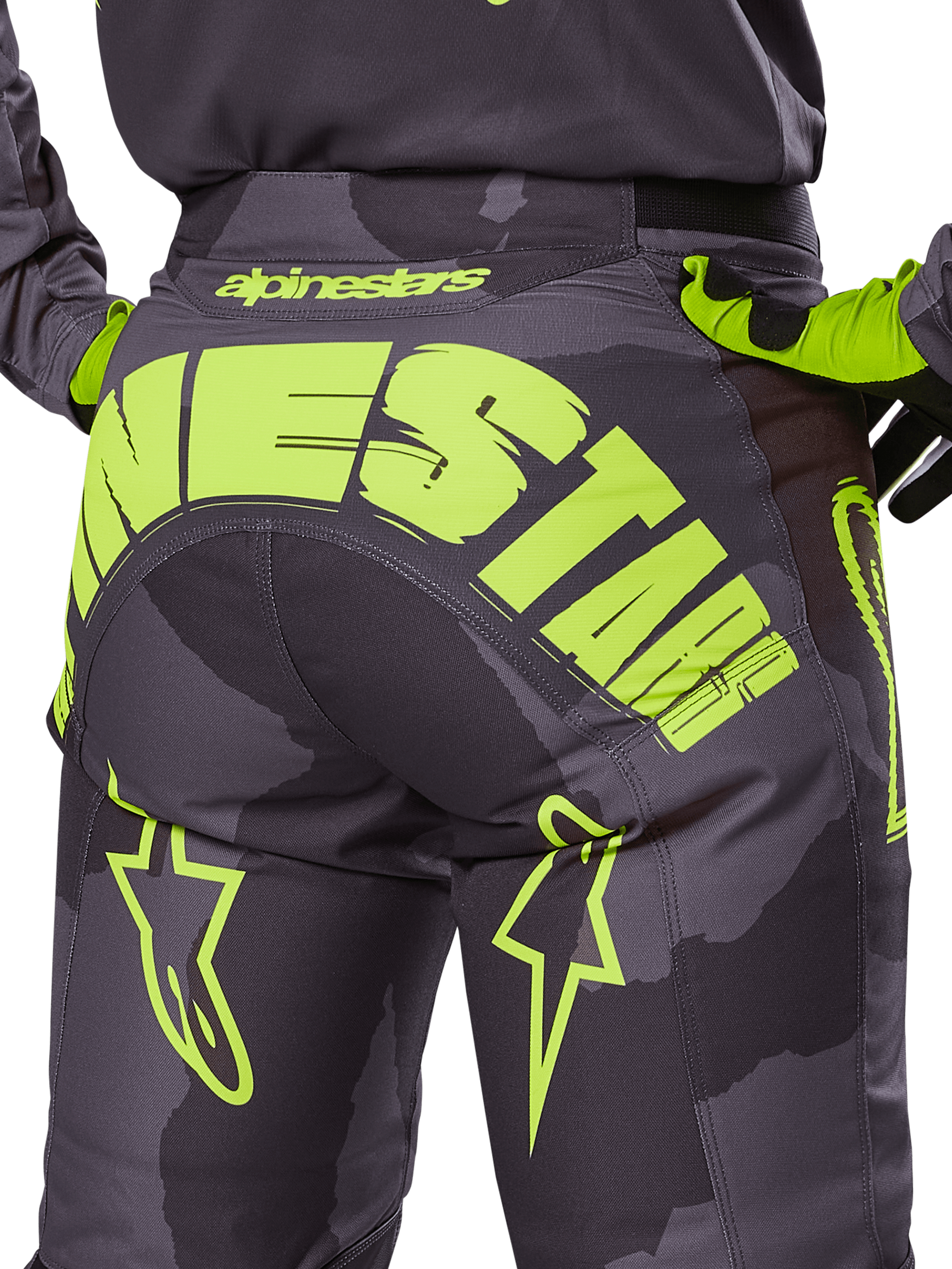 Alpinestars 2025 Racer Hollow Pants, spodnie motocrossowe, kolor ciemne camo i fluorescencyjny żółty, z dużym żółtym logo Alpinestars z tyłu, taktyczny wzór camo z jaskrawymi akcentami, wytrzymała konstrukcja do wyścigów terenowych