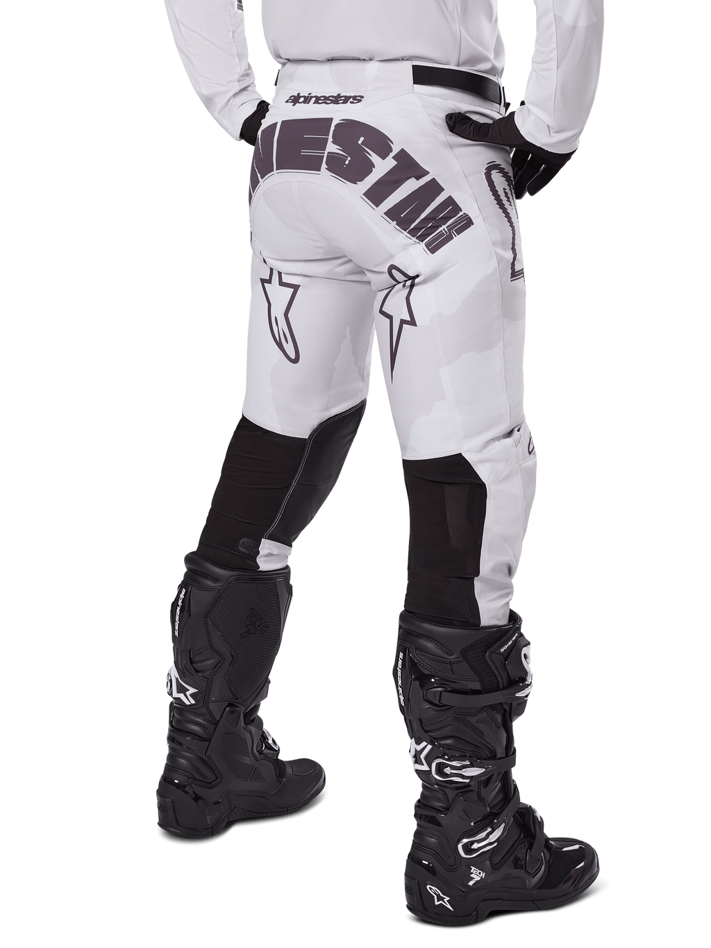 Alpinestars 2025 Racer Hollow Pants, spodnie motocrossowe, jasny brązowy kamuflaż, widok z tyłu na motocyklistę ukazujący duże logo na siedzeniu i czarne elastyczne panele na dolnej części nogawek, w połączeniu z czarnymi butami Tech 7