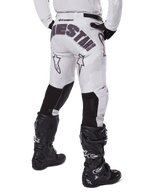 Alpinestars 2025 Racer Hollow Pants, spodnie motocrossowe, jasny brązowy kamuflaż, widok z tyłu na motocyklistę ukazujący duże logo na siedzeniu i czarne elastyczne panele na dolnej części nogawek, w połączeniu z czarnymi butami Tech 7