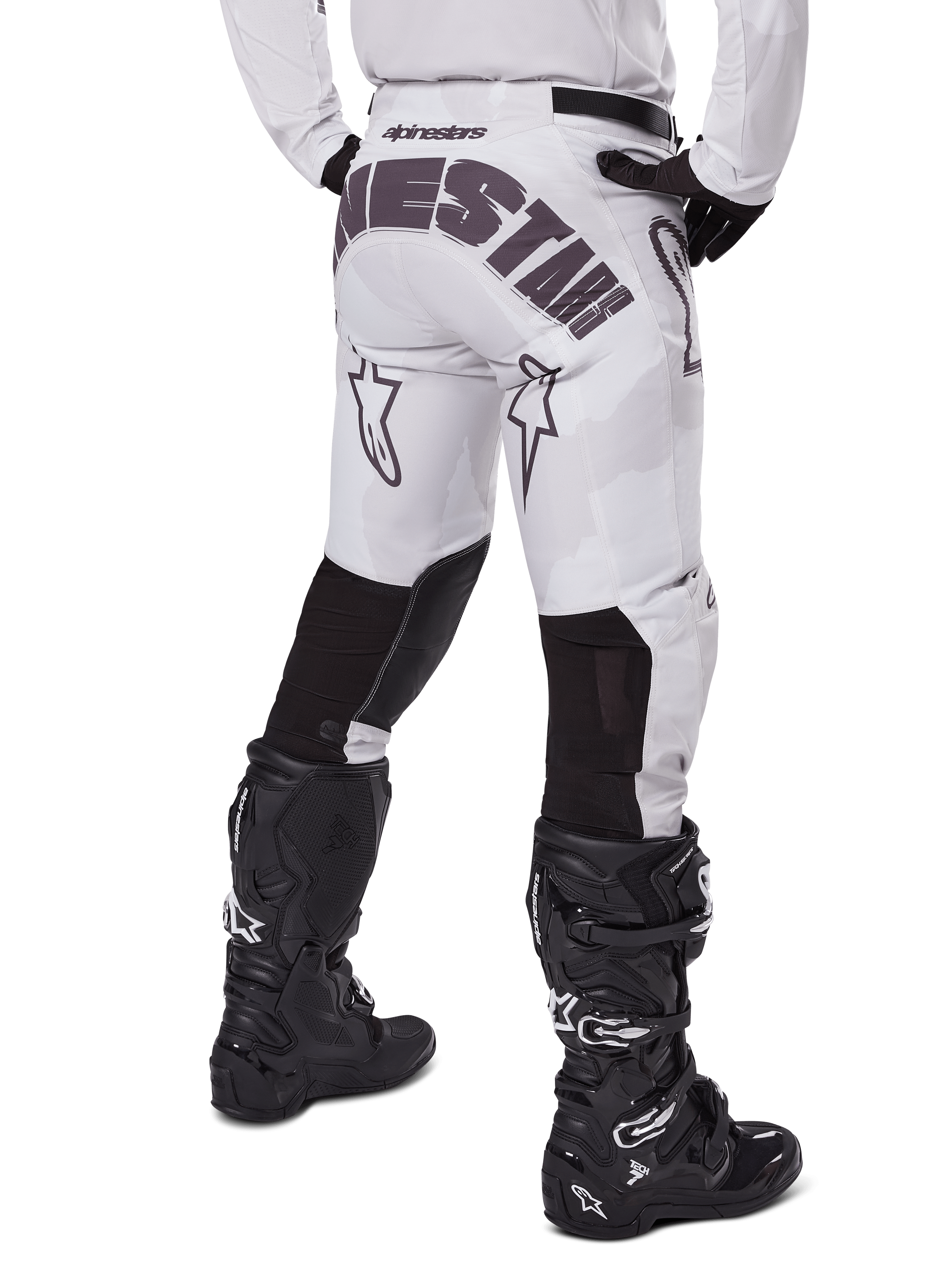 Alpinestars 2025 Racer Hollow Pants, spodnie motocrossowe, jasny brązowy kamuflaż, widok z tyłu na motocyklistę ukazujący duże logo na siedzeniu i czarne elastyczne panele na dolnej części nogawek, w połączeniu z czarnymi butami Tech 7
