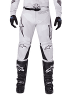 Alpinestars 2025 Racer Hollow Pants, spodnie motocrossowe, jasnobrązowy kamuflaż, widoczne z czarnymi rękawicami i butami motocrossowymi, posiadają subtelny wzór camo z czarnymi nadrukami graficznymi i logo Astars, ergonomiczny design do wyczynowej jazdy terenowej