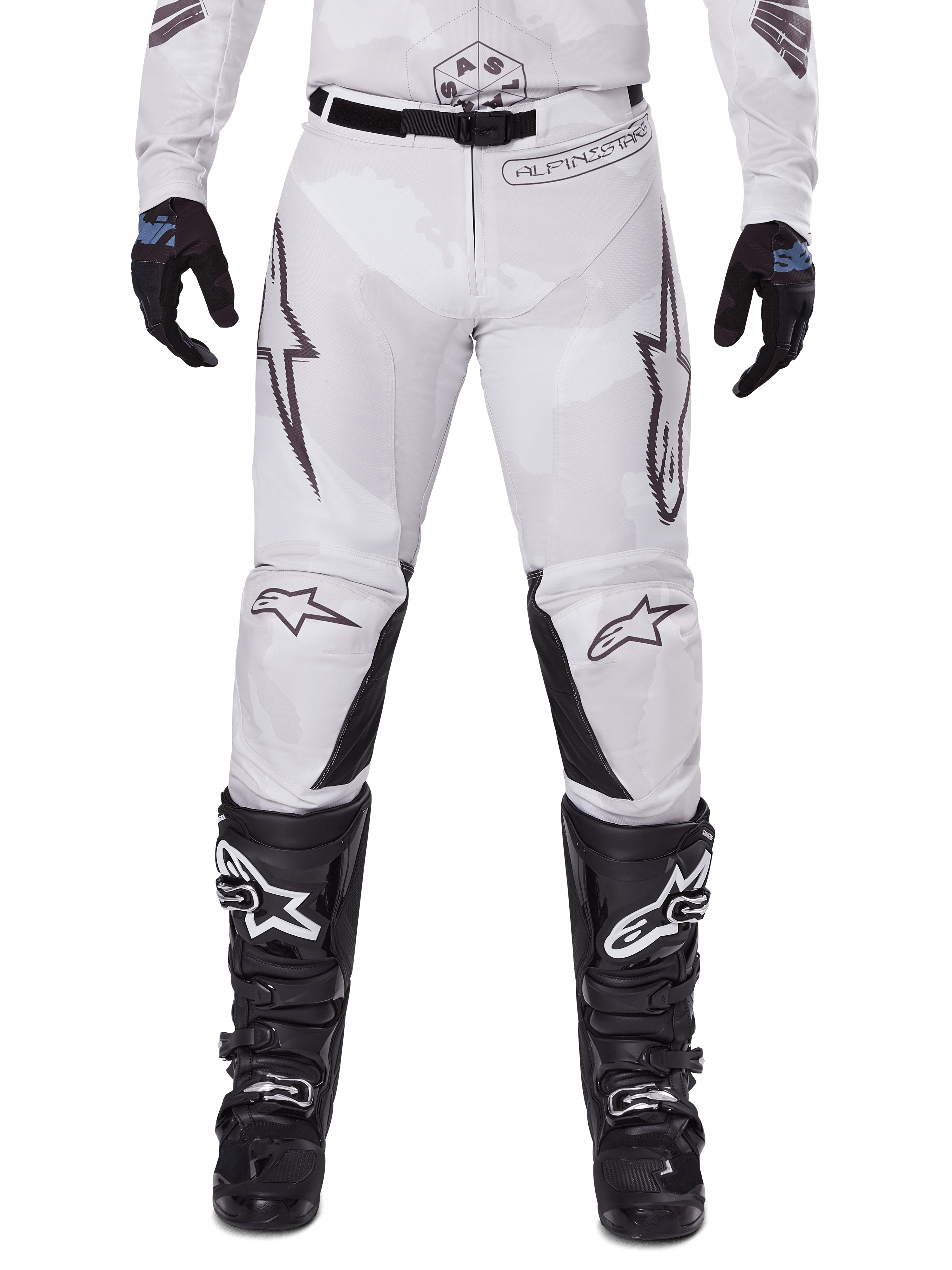 Alpinestars 2025 Racer Hollow Pants, spodnie motocrossowe, jasnobrązowy kamuflaż, widoczne z czarnymi rękawicami i butami motocrossowymi, posiadają subtelny wzór camo z czarnymi nadrukami graficznymi i logo Astars, ergonomiczny design do wyczynowej jazdy terenowej