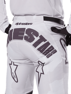 Widok z tyłu spodni Alpinestars 2025 Racer Hollow, spodnie motocrossowe, białe i jasny kamuflaż, z wyraźnym czarnym logo Alpinestars na siedzeniu, noszone z pasującą koszulką i czarnymi rękawicami