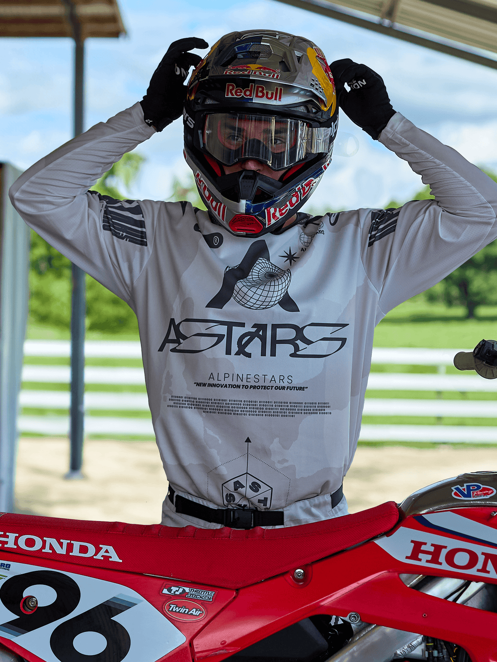 Zawodnik motocrossowy w stroju Alpinestars 2025 Racer Hollow, koszulka w jasnym kamuflażu z wyraźnym brandingiem ASTARS, poprawiający kask Red Bull z przezroczystymi goglami, stojący obok czerwonego motocykla Honda w scenerii terenowej