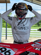 Zawodnik motocrossowy w stroju Alpinestars 2025 Racer Hollow, koszulka w jasnym kamuflażu z wyraźnym brandingiem ASTARS, poprawiający kask Red Bull z przezroczystymi goglami, stojący obok czerwonego motocykla Honda w scenerii terenowej