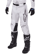 Alpinestars Racer Hollow Pants 2025, spodnie motocrossowe, biało-szary kamuflaż, z dużymi czarnymi konturowymi logo gwiazdy i czarnymi skórzanymi panelami po wewnętrznej stronie kolan, noszone z czarnymi butami i rękawicami motocrossowymi, wytrzymała konstrukcja do wyścigów terenowych.