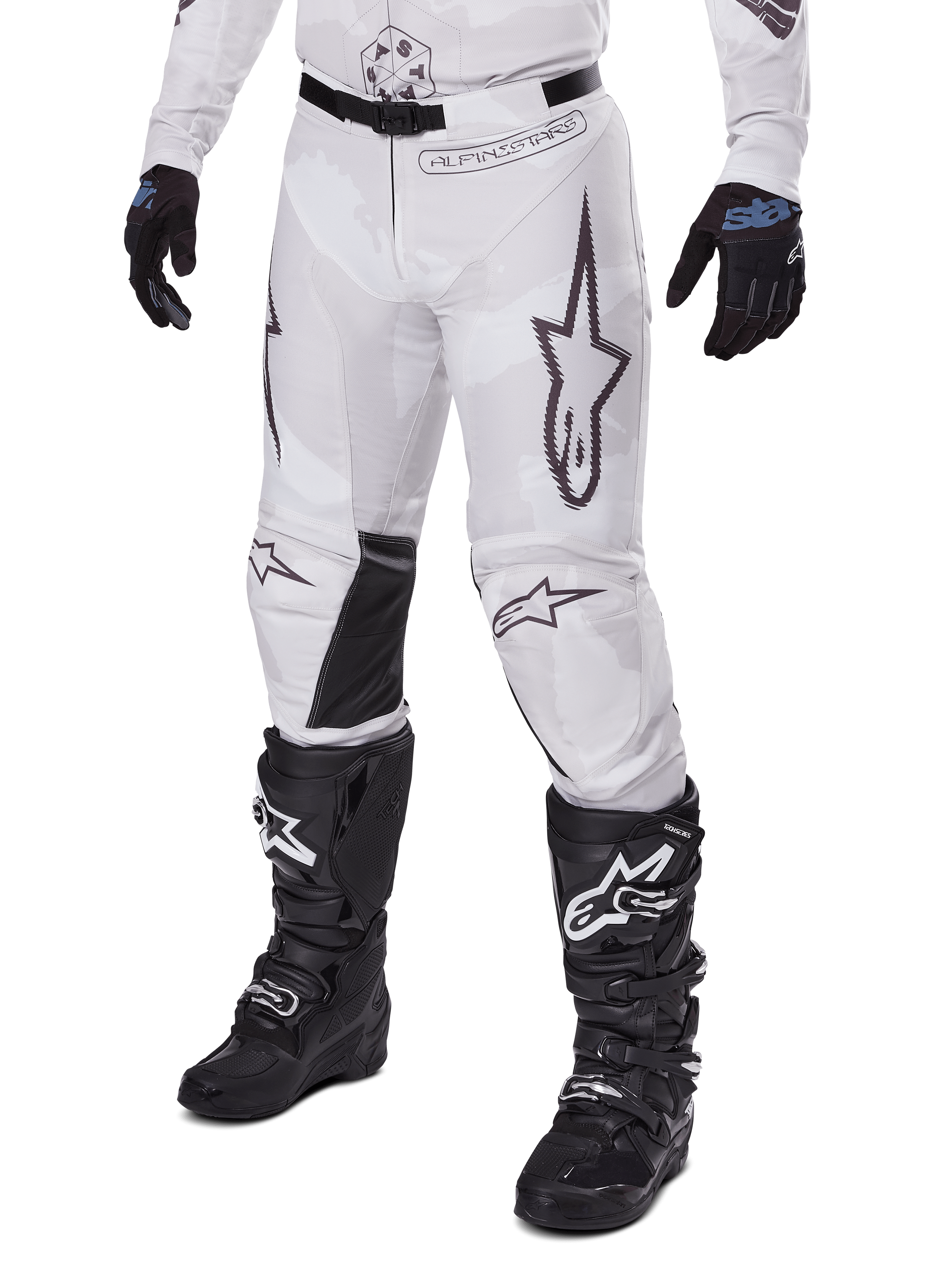 Alpinestars Racer Hollow Pants 2025, spodnie motocrossowe, biało-szary kamuflaż, z dużymi czarnymi konturowymi logo gwiazdy i czarnymi skórzanymi panelami po wewnętrznej stronie kolan, noszone z czarnymi butami i rękawicami motocrossowymi, wytrzymała konstrukcja do wyścigów terenowych.