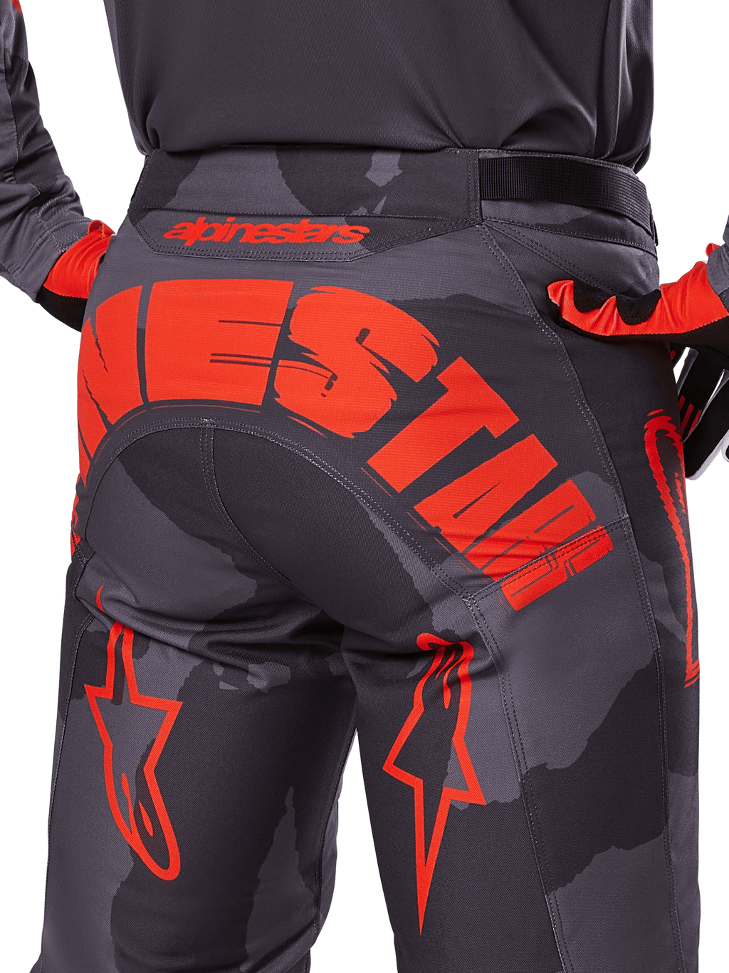Alpinestars 2025 Racer Hollow Pants, spodnie motocrossowe, szary kamuflaż z jaskrawym pomarańczowym, widok z tyłu pokazujący duże pomarańczowe logo Alpinestars na siedzeniu, wzór kamuflażu z neonowymi akcentami, wytrzymała odzież do jazdy w terenie