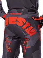 Alpinestars 2025 Racer Hollow Pants, spodnie motocrossowe, szary kamuflaż z jaskrawym pomarańczowym, widok z tyłu pokazujący duże pomarańczowe logo Alpinestars na siedzeniu, wzór kamuflażu z neonowymi akcentami, wytrzymała odzież do jazdy w terenie