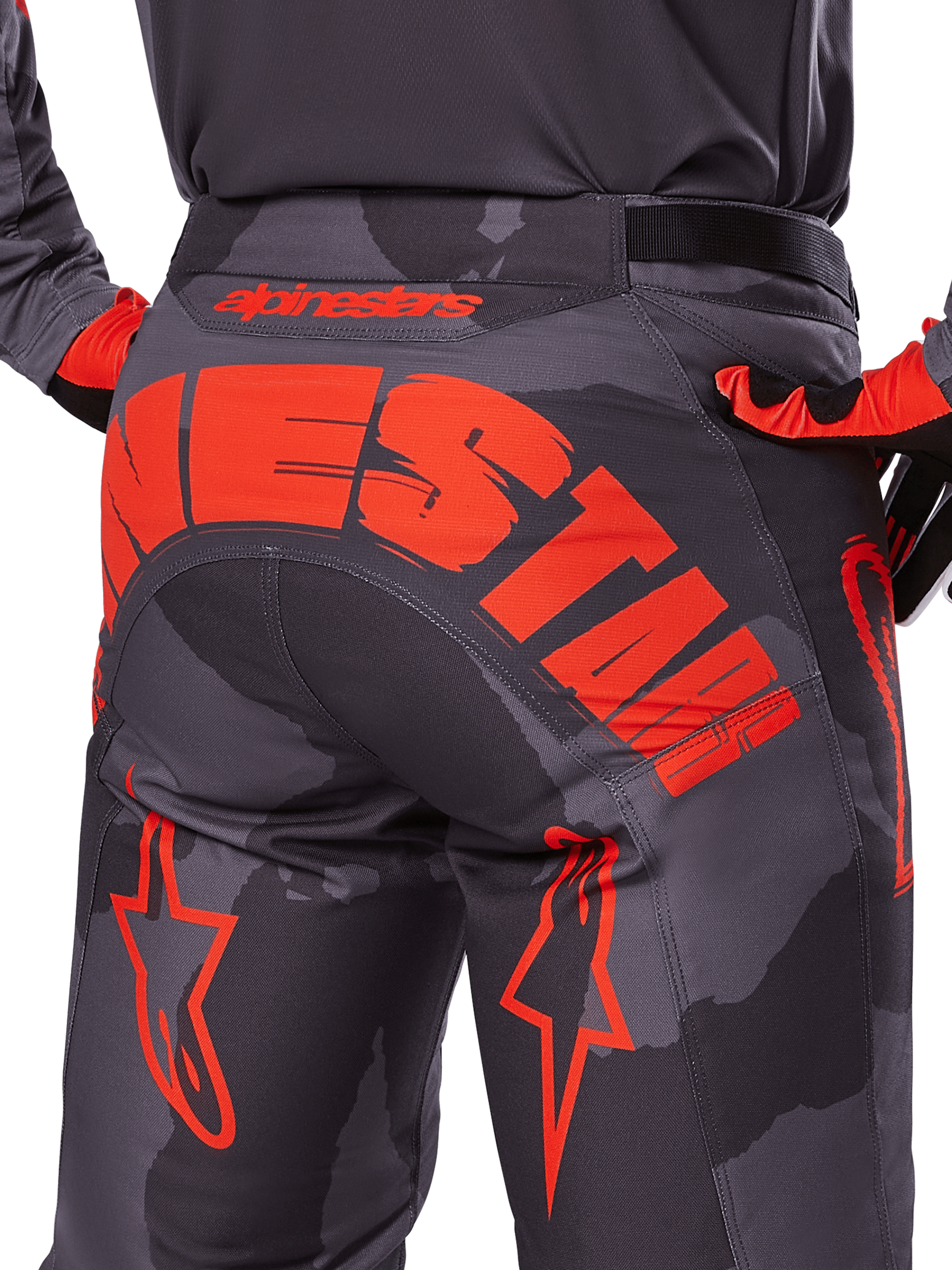 Alpinestars 2025 Racer Hollow Pants, spodnie motocrossowe, szary kamuflaż z jaskrawym pomarańczowym, widok z tyłu pokazujący duże pomarańczowe logo Alpinestars na siedzeniu, wzór kamuflażu z neonowymi akcentami, wytrzymała odzież do jazdy w terenie