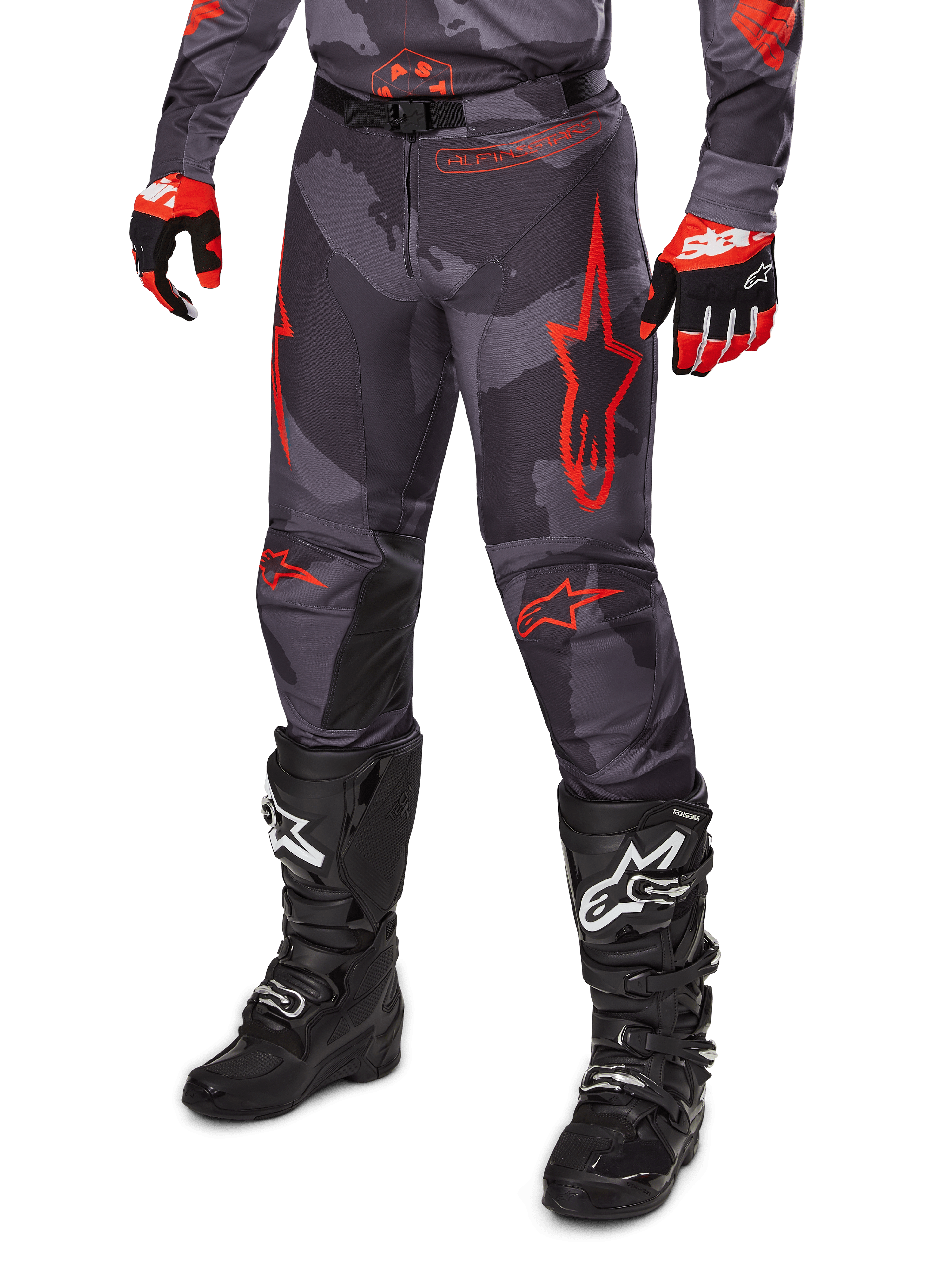 Alpinestars 2025 Racer Hollow Pants, spodnie motocrossowe, szare moro i czerwone, noszone z czarnymi butami i czerwonymi rękawicami, wyraziste czerwone logo w kształcie gwiazdy, wzór kamuflażu i wytrzymałe panele ochronne do jazdy terenowej