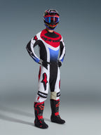 Alpinestars 2026 Techstar Knif Pants, spodnie motocrossowe, czarno-biało-czerwone, prezentowane na zawodniku w pełnym stroju wyścigowym z pasującą bluzą i butami, z wyrazistymi czerwonymi logo Alpinestars na białych panelach, o sportowym kroju.