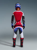 Alpinestars 2026 Techstar Knif Pants, spodnie motocrossowe, czarne, białe, czerwone i niebieskie, widok od tyłu zawodnika w pełnym stroju, w tym pasująca koszulka, niebieski kask oraz czarno-czerwone buty, z wyraźnym brandingiem Alpinestars na siedzeniu.