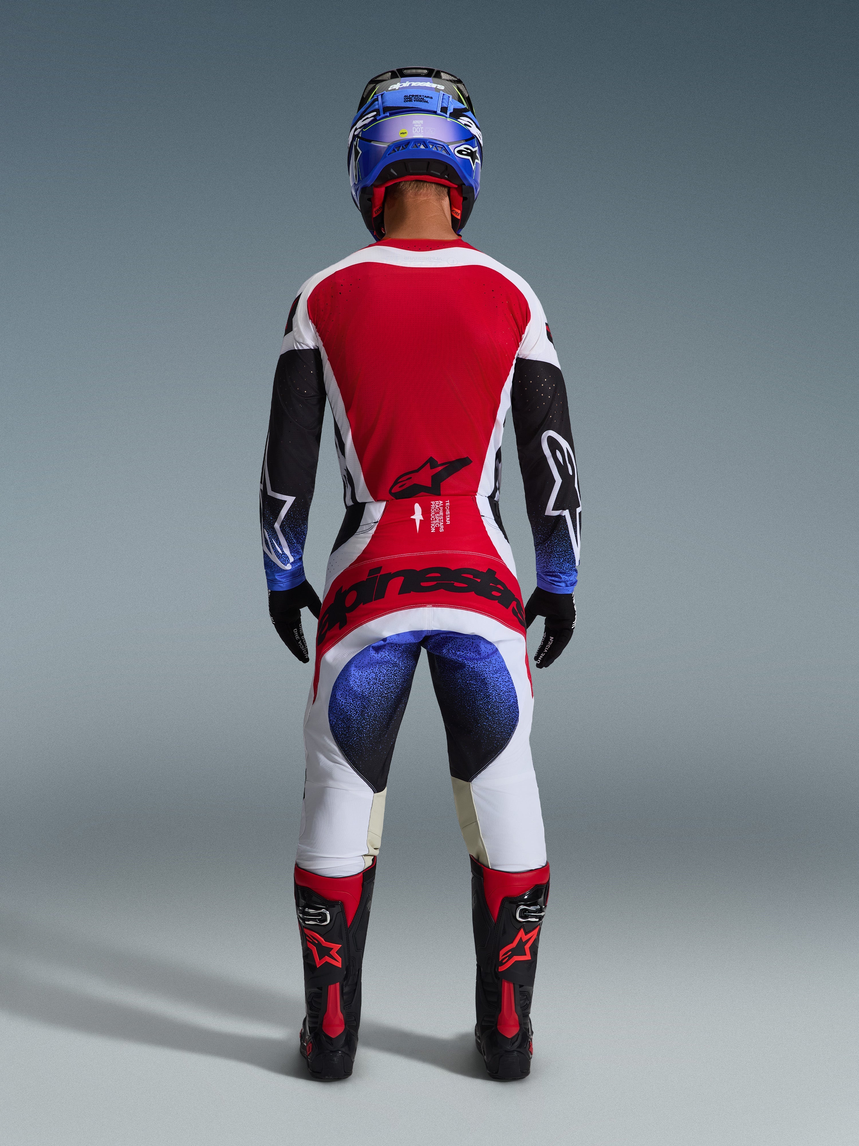 Alpinestars 2026 Techstar Knif Pants, spodnie motocrossowe, czarne, białe, czerwone i niebieskie, widok od tyłu zawodnika w pełnym stroju, w tym pasująca koszulka, niebieski kask oraz czarno-czerwone buty, z wyraźnym brandingiem Alpinestars na siedzeniu.