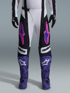 Alpinestars 2026 Techstar Knif Pants, spodnie motocrossowe, czarne, białe i fioletowe z różowymi i niebieskimi akcentami, noszone z fioletowymi butami wyścigowymi i czarnymi rękawicami, charakteryzujące się ergonomicznym krojem wyścigowym i wyrazistymi grafikami logo dla wydajności w terenie