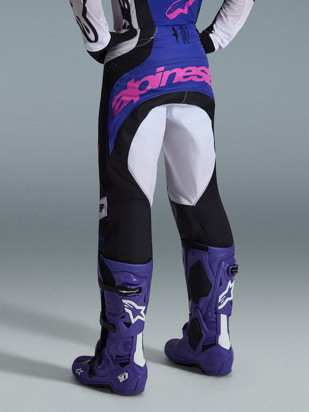 Spodnie Alpinestars Techstar Knif 2026, spodnie motocrossowe, czarne, jasnoszare i fioletowe, widok z tyłu z różowym logotypem na siedzeniu, noszone z dopasowaną koszulką i fioletowymi butami motocrossowymi, profesjonalny strój wyścigowy off-road