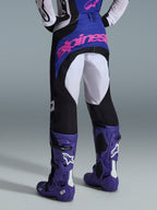 Spodnie Alpinestars Techstar Knif 2026, spodnie motocrossowe, czarne, jasnoszare i fioletowe, widok z tyłu z różowym logotypem na siedzeniu, noszone z dopasowaną koszulką i fioletowymi butami motocrossowymi, profesjonalny strój wyścigowy off-road