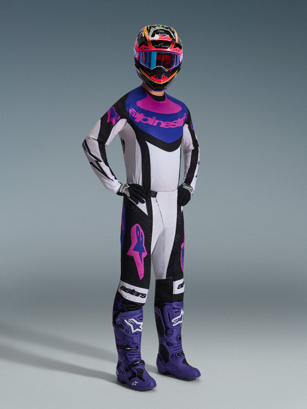 Zawodnik motocrossowy w zestawie Alpinestars 2026 Techstar Knif, biała koszulka z czarnymi, niebieskimi i różowymi akcentami, dopasowane spodnie z różowymi logotypami Astars, wielobarwny kask z goglami, fioletowe buty wyścigowe, profesjonalna odzież wyczynowa do motocrossu
