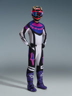 Motocyklista motocrossowy w zestawie Alpinestars 2026 Techstar Knif, kolor czarny, jasnoszary i fioletowy, biała koszulka z niebieskimi i różowymi akcentami, pasujące spodnie z fioletowymi logo gwiazdy, kolorowy różowo-żółty kask, fioletowe buty i czarne rękawice.