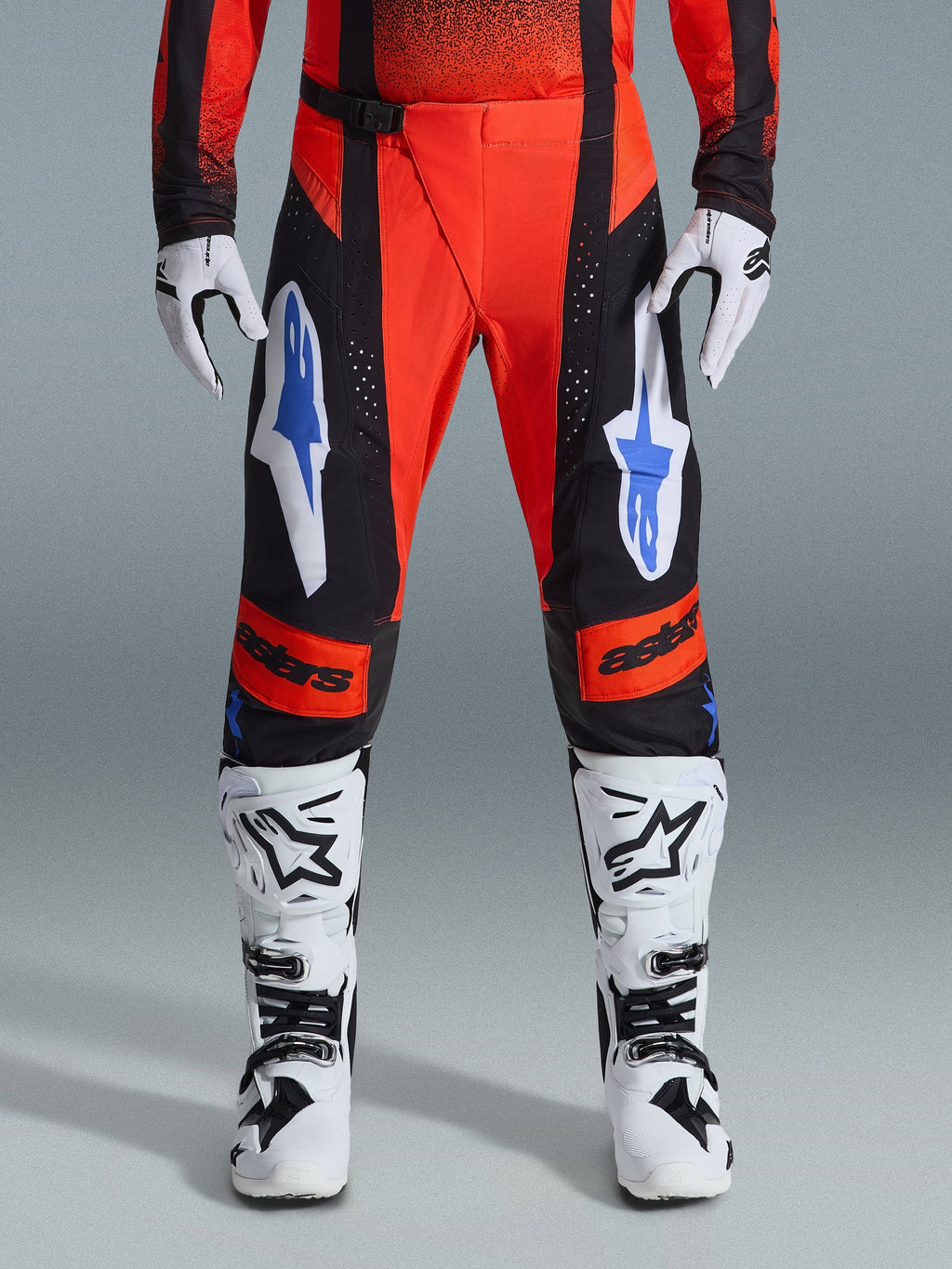 Alpinestars 2026 Techstar Knif Pants, spodnie motocrossowe, pomarańczowe, czarne i niebieskie UCLA Blue, noszone z dopasowaną koszulką, biało-czarnymi butami motocrossowymi i rękawicami, z wyrazistym brandingiem na udach i kolanach, sportowy krój wyścigowy do jazdy off-road