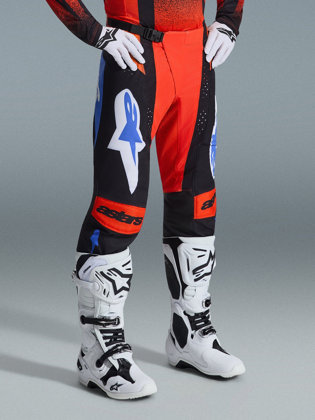 Alpinestars Techstar Knif Pants 2026, spodnie motocrossowe, pomarańczowe, czarne i niebieskie (UCLA Blue), zestawione z białymi rękawicami i biało-czarnymi butami motocrossowymi, wyraźne logo na udach i pomarańczowe panele na kolanach z logo astars
