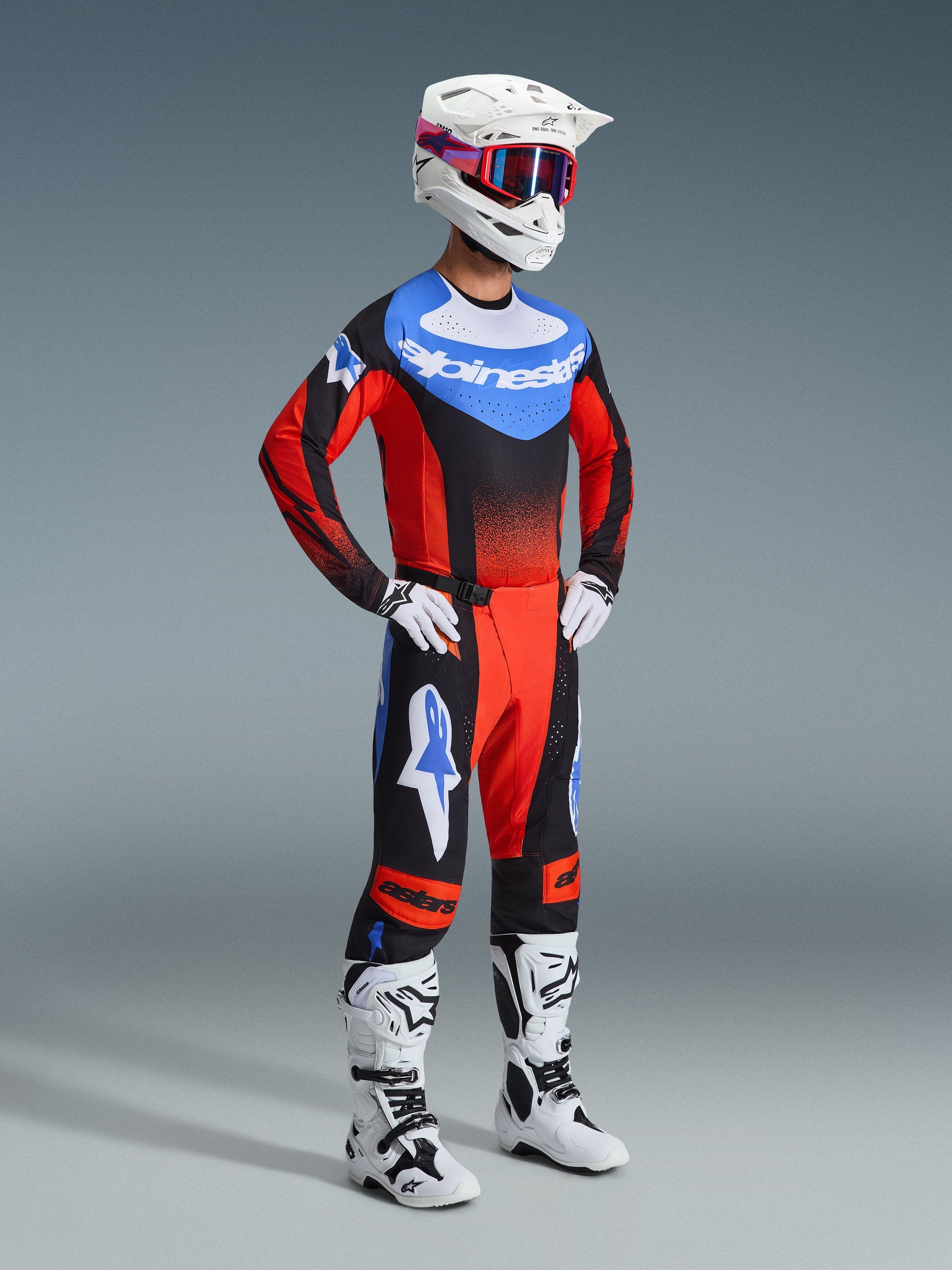 Zawodnik motocrossowy w spodniach Techstar Knif 2026 w kolorach pomarańczowym, czarnym i UCLA Blue, dopasowana koszulka wyścigowa, biały kask z czerwono-niebieskimi goglami, białe buty off-road, kompletny profesjonalny zestaw odzieży motocrossowej.