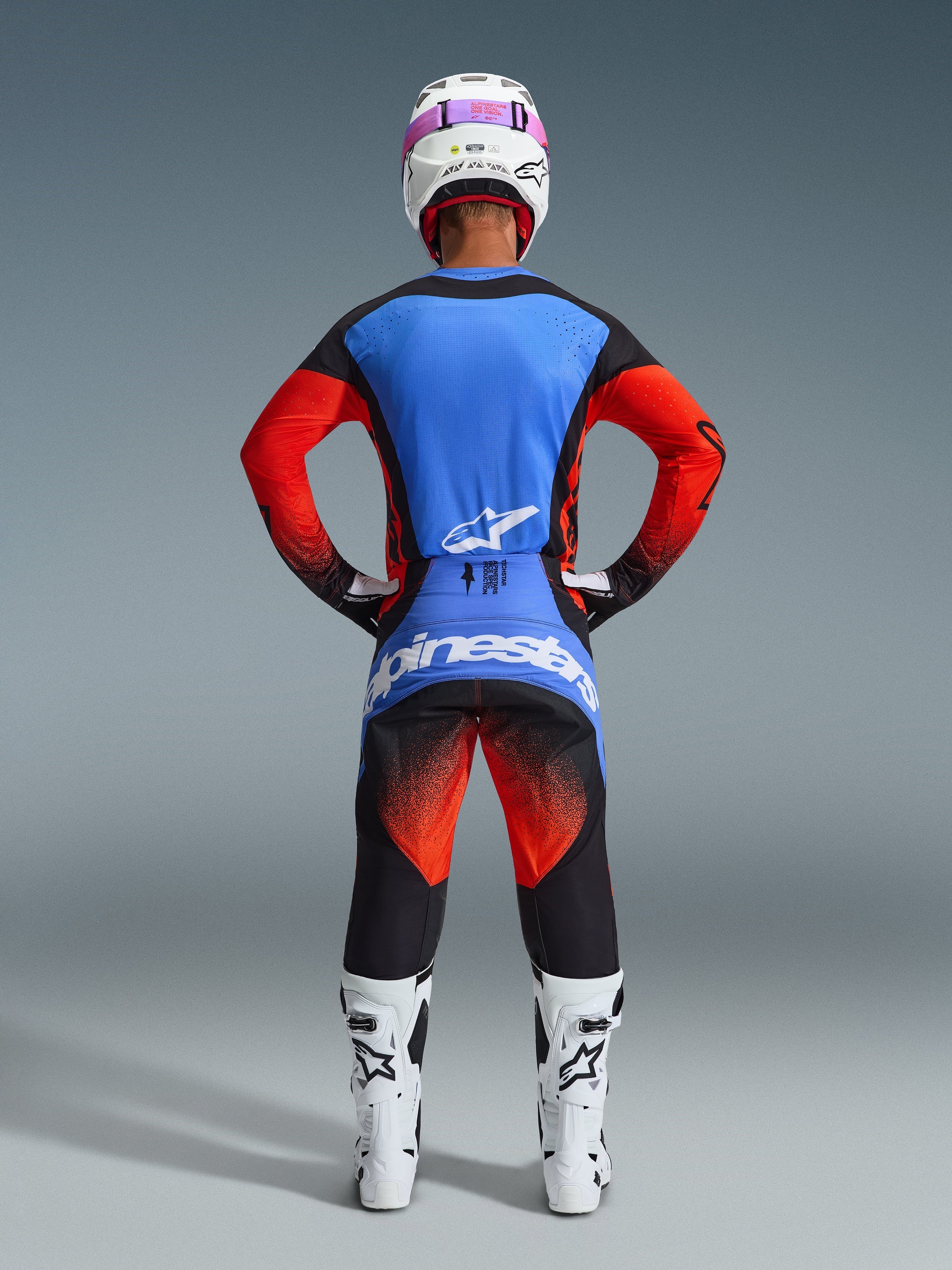 Zawodnik motocrossowy widziany od tyłu, ubrany w spodnie Techstar Knif 2026 w kolorze pomarańczowym, czarnym i UCLA Blue, zestawione z niebiesko-pomarańczową koszulką, białym kaskiem z różowymi goglami i białymi butami wyścigowymi, pełny strój Alpinestars do zawodów off-road
