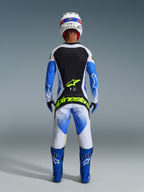 Motocyklista motocrossowy widoczny od tyłu w pełnym stroju wyścigowym, spodnie Techstar Knif 2026 w kolorze UCLA Blue Black i Yellow Fluo, pasująca koszulka z długim rękawem z czarno-białymi panelami, niebieskie buty off-road, biały kask, z wyrazistym neonowożółtym logo Alpinestars na tyle spodni.