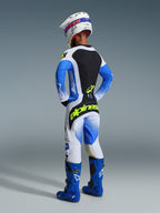 Zawodnik motocrossowy widziany od tyłu, ubrany w spodnie 2026 Techstar Knif w kolorze Ucla Blue Black i Yellow Fluo, z jaskrawożółtym logo na siedzeniu, dopasowaną koszulką z niebieskimi rękawami, białym kaskiem i niebieskimi butami off-road.