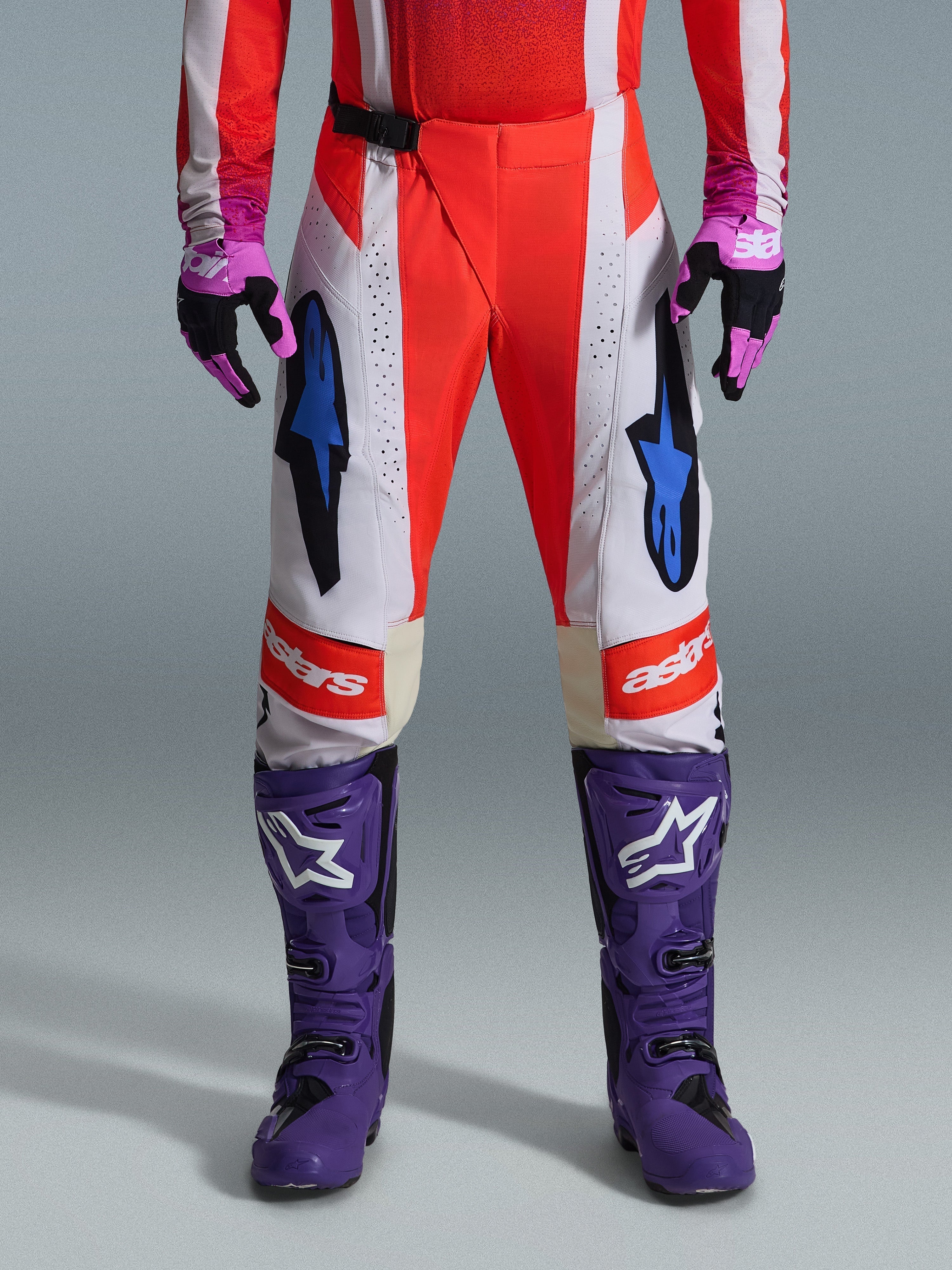 Alpinestars 2026 Techstar Knif Pants, spodnie motocrossowe, pomarańczowe fluo i jasnoszare z czarnymi akcentami, widoczne z fioletowymi butami i rękawicami, niebieskie logo gwiazdy, wielopanelowa konstrukcja zapewniająca wydajność w terenie