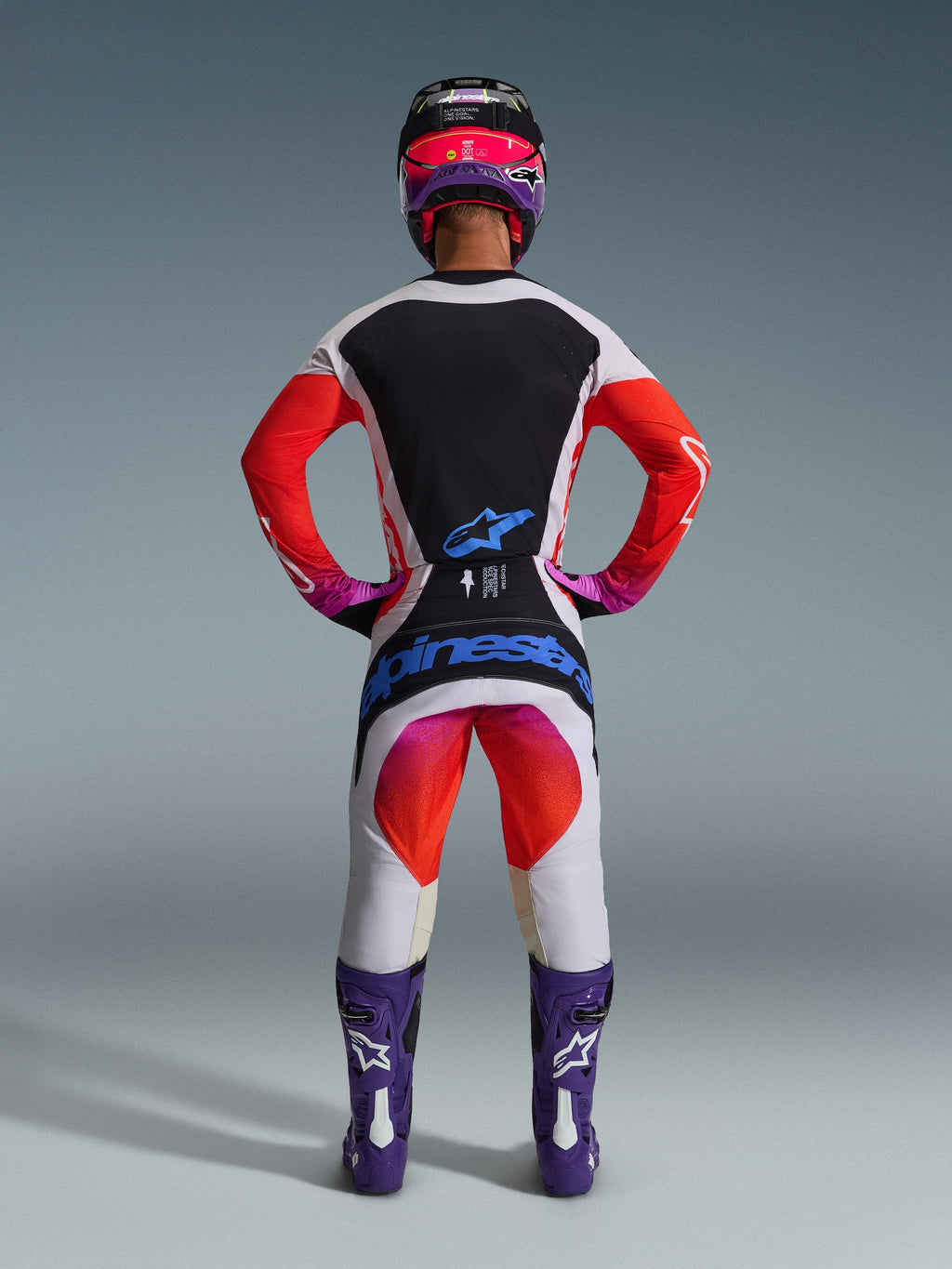 Motocyklista motocrossowy widziany od tyłu w spodniach Techstar Knif 2026 i pasującej bluzie, jasnoszaro-czarny design z jaskrawymi pomarańczowymi i fioletowymi akcentami, niebieskie logo Alpinestars na siedzeniu, fioletowe buty i kask, profesjonalny zestaw odzieży do wyścigów terenowych