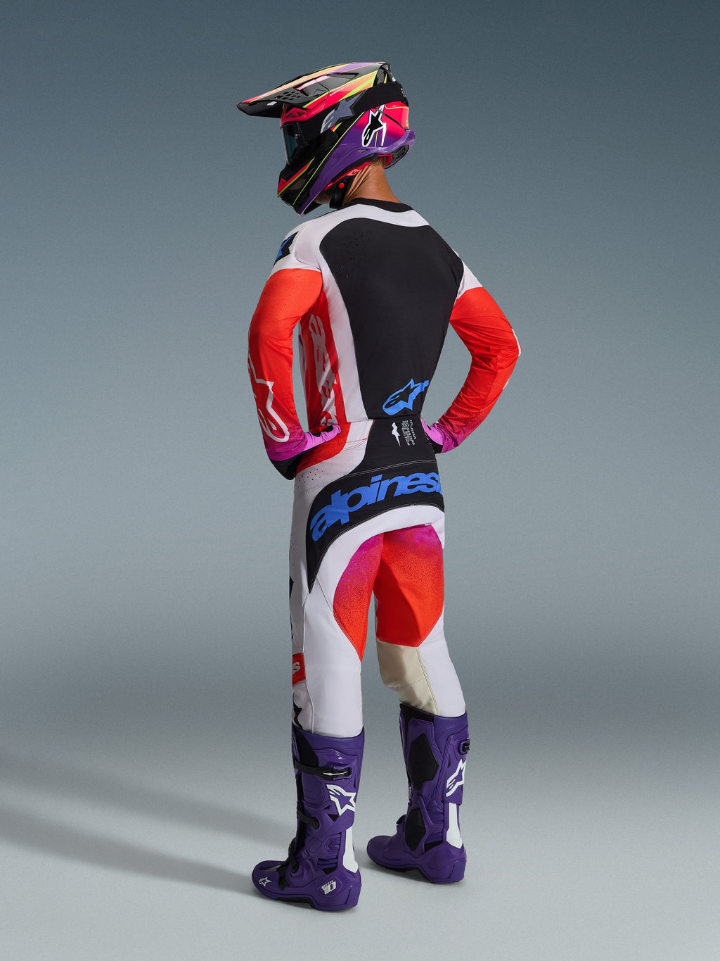 Motocyklista motocrossowy widziany od tyłu w pełnym stroju wyścigowym, spodnie Alpinestars 2026 Techstar Knif w kolorze jasnoszarym i jaskrawopomarańczowym (Orange Fluo) z niebieskimi oznaczeniami, dopasowana koszulka z pomarańczowymi rękawami, fioletowe buty Tech 10 oraz kolorowy kask wyścigowy, profesjonalny zestaw off-road