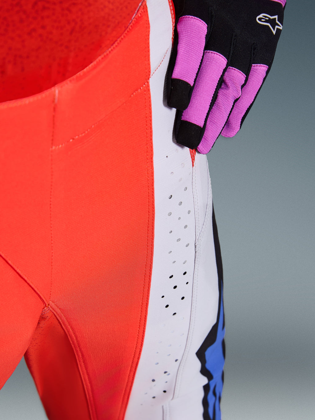 Alpinestars Techstar Knif Pants 2026, spodnie motocrossowe, pomarańczowy fluo i jasnoszary z niebieskimi akcentami, zbliżenie pokazujące laserowo wycinane perforacje wentylacyjne na udzie, widoczna czarno-fioletowa rękawica motocrossowa