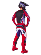 Motocyklista motocrossowy widziany z tyłu w spodniach Alpinestars 2025 Fluid Wurx, fioletowo-czerwonych z białymi i czarnymi akcentami, pasująca koszulka, czarny kask i czerwone buty, dopasowany strój do wyścigów terenowych