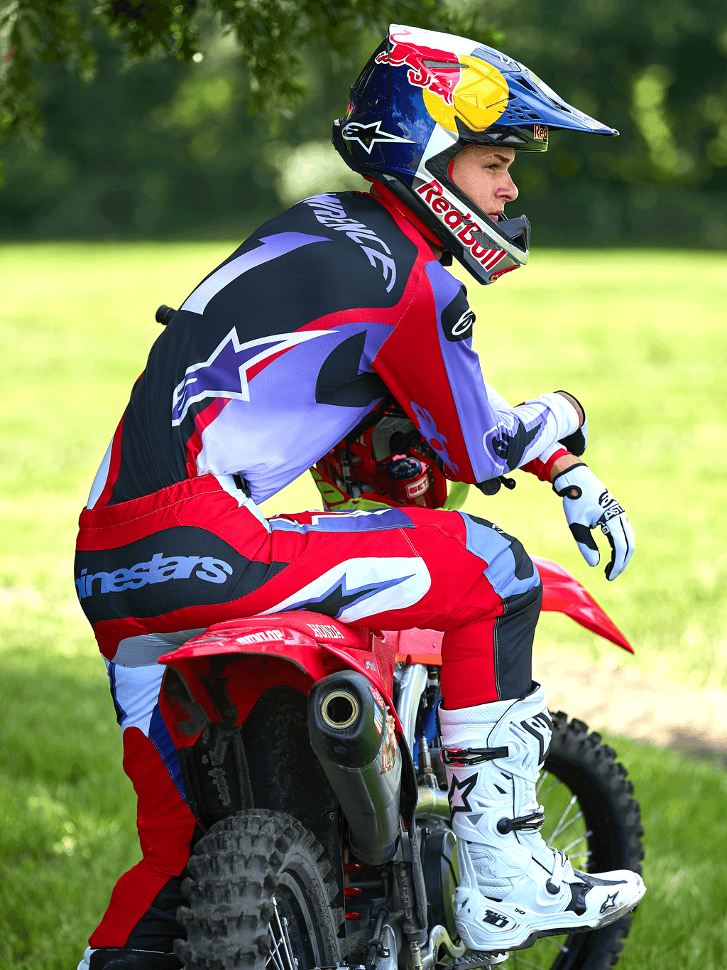 Motocrossista siedzący na czerwonym motocyklu crossowym, ubrany w spodnie Alpinestars 2025 Fluid Wurx w kolorze fioletowym i jaskrawoczerwonym, pasującą koszulkę, biało-czarne buty Tech 10, białe rękawice i kask Red Bull, kompletny strój do wyścigów terenowych w plenerze