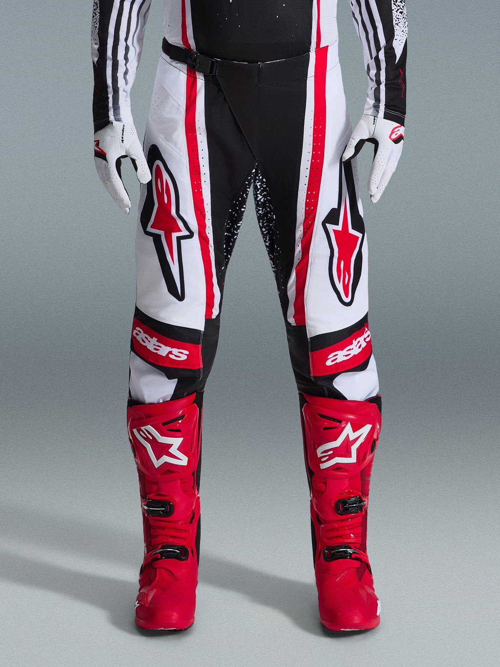 Alpinestars 2026 Techstar Nomur Pants, spodnie motocrossowe, czarno-biało-czerwone, noszone z dopasowanymi czerwonymi butami i białymi rękawicami, z wyraźnymi czerwonymi logo na udach i brandingiem „astars” na kolanach, wielopanelowa konstrukcja wyścigowa do jazdy w terenie
