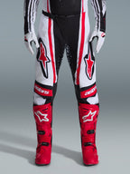 Alpinestars 2026 Techstar Nomur Pants, spodnie motocrossowe, czarno-biało-czerwone, noszone z dopasowanymi czerwonymi butami i białymi rękawicami, z wyraźnymi czerwonymi logo na udach i brandingiem „astars” na kolanach, wielopanelowa konstrukcja wyścigowa do jazdy w terenie
