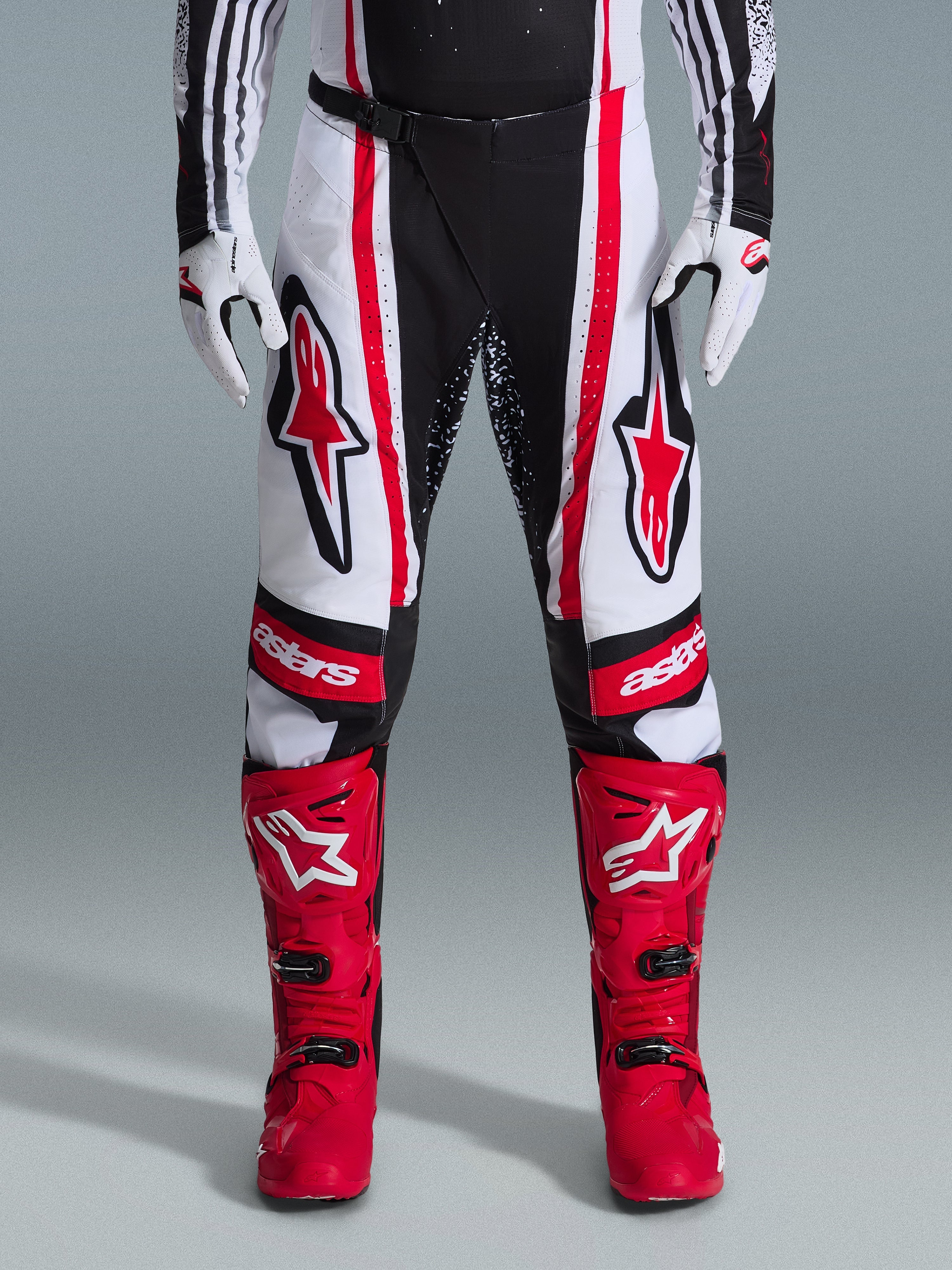 Alpinestars 2026 Techstar Nomur Pants, spodnie motocrossowe, czarno-biało-czerwone, noszone z dopasowanymi czerwonymi butami i białymi rękawicami, z wyraźnymi czerwonymi logo na udach i brandingiem „astars” na kolanach, wielopanelowa konstrukcja wyścigowa do jazdy w terenie