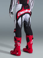 Alpinestars Techstar Nomur Pants 2026, spodnie motocrossowe, czarno-biało-czerwone, widok z tyłu na motocykliście w czerwonych butach, charakterystyczne logo „astars” na siedzeniu i nakrapiane wzory, profesjonalny sprzęt off-road