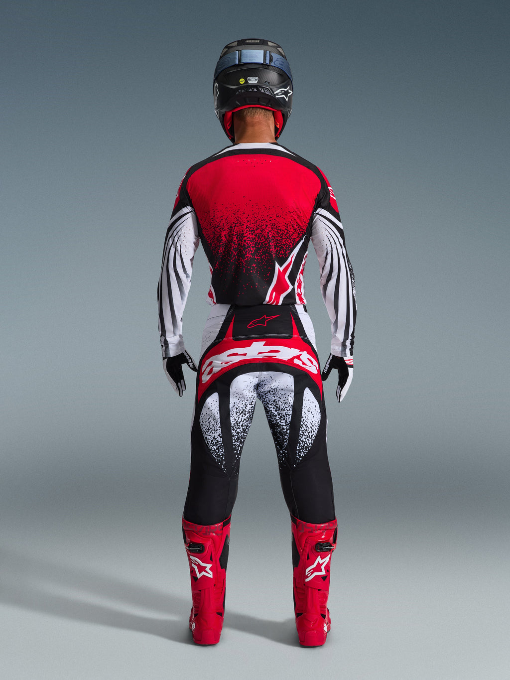 Widok z tyłu zawodnika motocrossowego ubranego w spodnie Alpinestars 2026 Techstar Nomur w kolorze czarno-biało-czerwonym, z wyraźnym logo na siedzeniu i nakrapianą grafiką, w zestawieniu z pasującą czerwoną koszulką i jaskrawoczerwonymi butami, tworzących pełny strój do wyścigów terenowych.