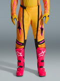 Alpinestars 2026 Techstar Nomur Pants, spodnie motocrossowe, pomarańczowo-żółto-różowe, noszone z pasującymi żółtymi rękawicami i jaskraworóżowymi butami wyścigowymi, czarne panele i wyraźne różowe logo Alpinestars, techniczna odzież wyścigowa off-road