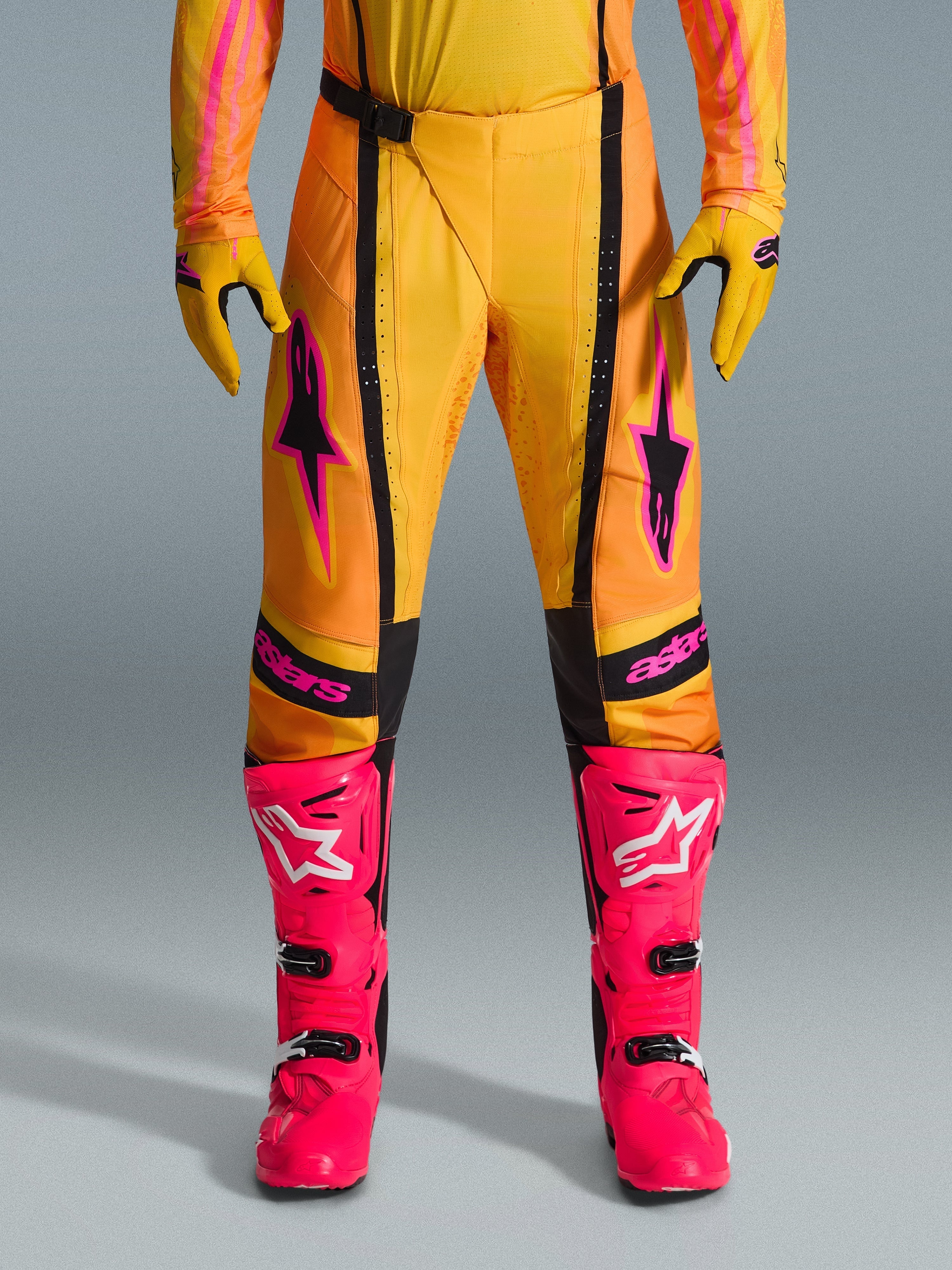 Alpinestars 2026 Techstar Nomur Pants, spodnie motocrossowe, pomarańczowo-żółto-różowe, noszone z pasującymi żółtymi rękawicami i jaskraworóżowymi butami wyścigowymi, czarne panele i wyraźne różowe logo Alpinestars, techniczna odzież wyścigowa off-road