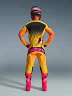 Widok z tyłu zawodnika motocrossowego ubranego w spodnie Alpinestars 2026 Techstar Nomur w kolorze pomarańczowo-żółto-różowym, z dużym różowym logo na siedzeniu, w połączeniu z dopasowaną żółto-czarną koszulką, jaskraworóżowymi butami i czarno-różowym kaskiem.