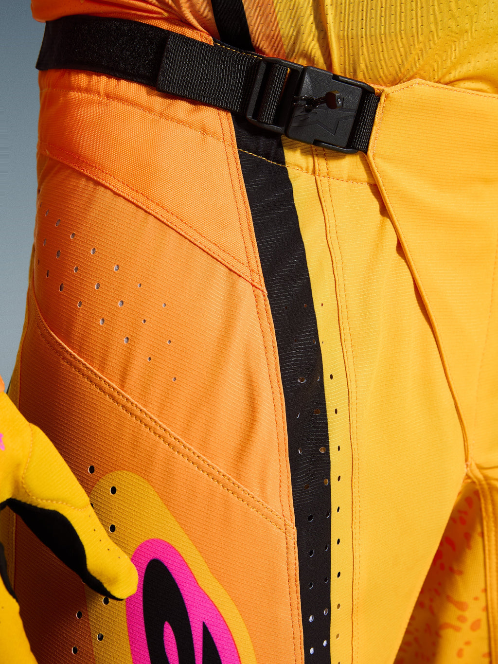 Alpinestars 2026 Techstar Nomur Pants, spodnie motocrossowe, pomarańczowo-żółto-różowe, zbliżenie na okolicę pasa z magnetycznym zapięciem na klamrę SX2, czarnym paskiem regulacyjnym i laserowo wycinanymi perforacjami wentylacyjnymi na wytrzymałym materiale ripstop