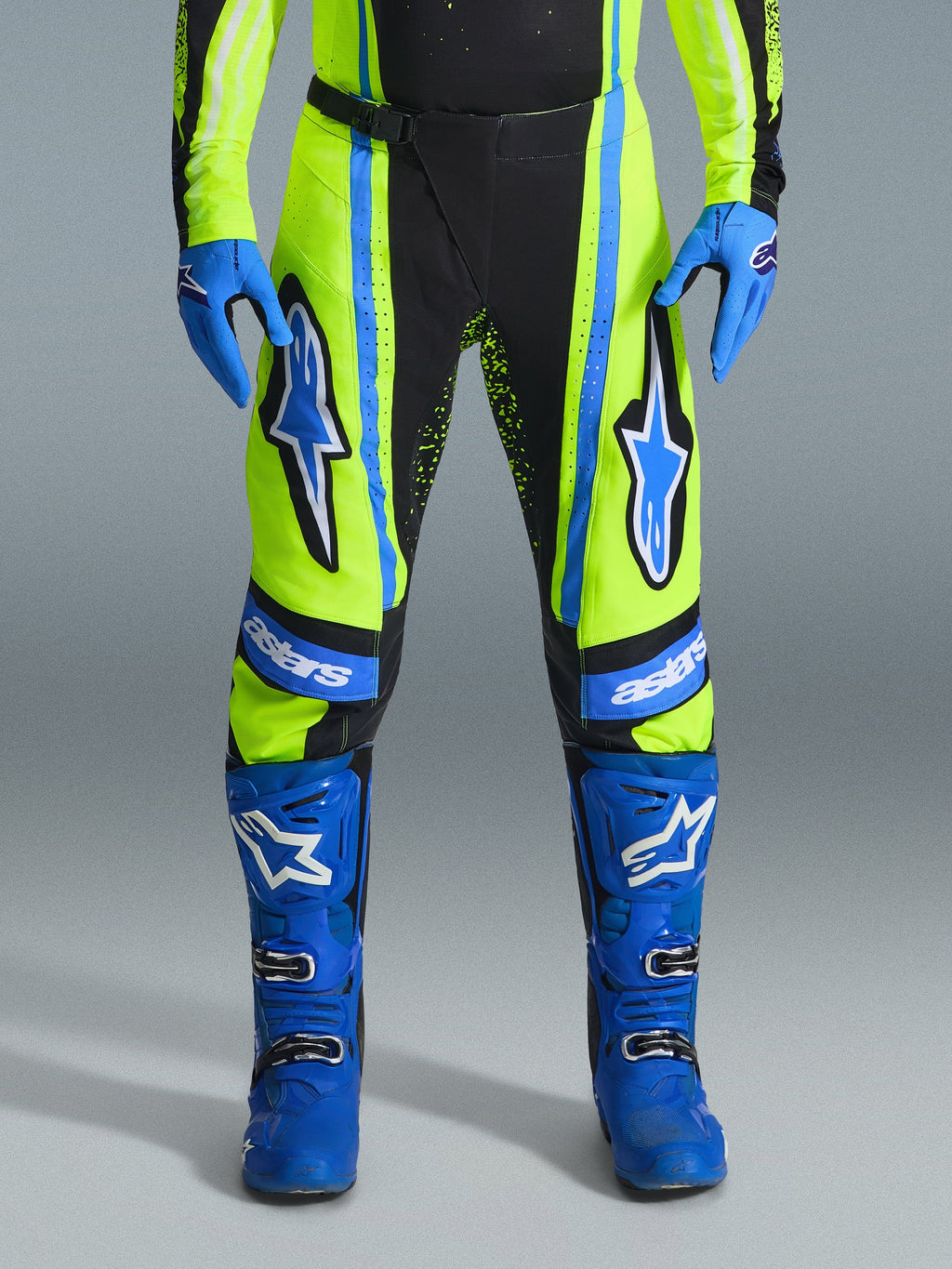Alpinestars 2026 Techstar Nomur Pants, spodnie motocrossowe, żółte neonowe (fluo) i ciemnogranatowe z niebieskimi akcentami, noszone z dopasowanymi niebieskimi rękawicami i butami, wyraźne logo Astars na udach, wyczynowa konstrukcja do wyścigów terenowych